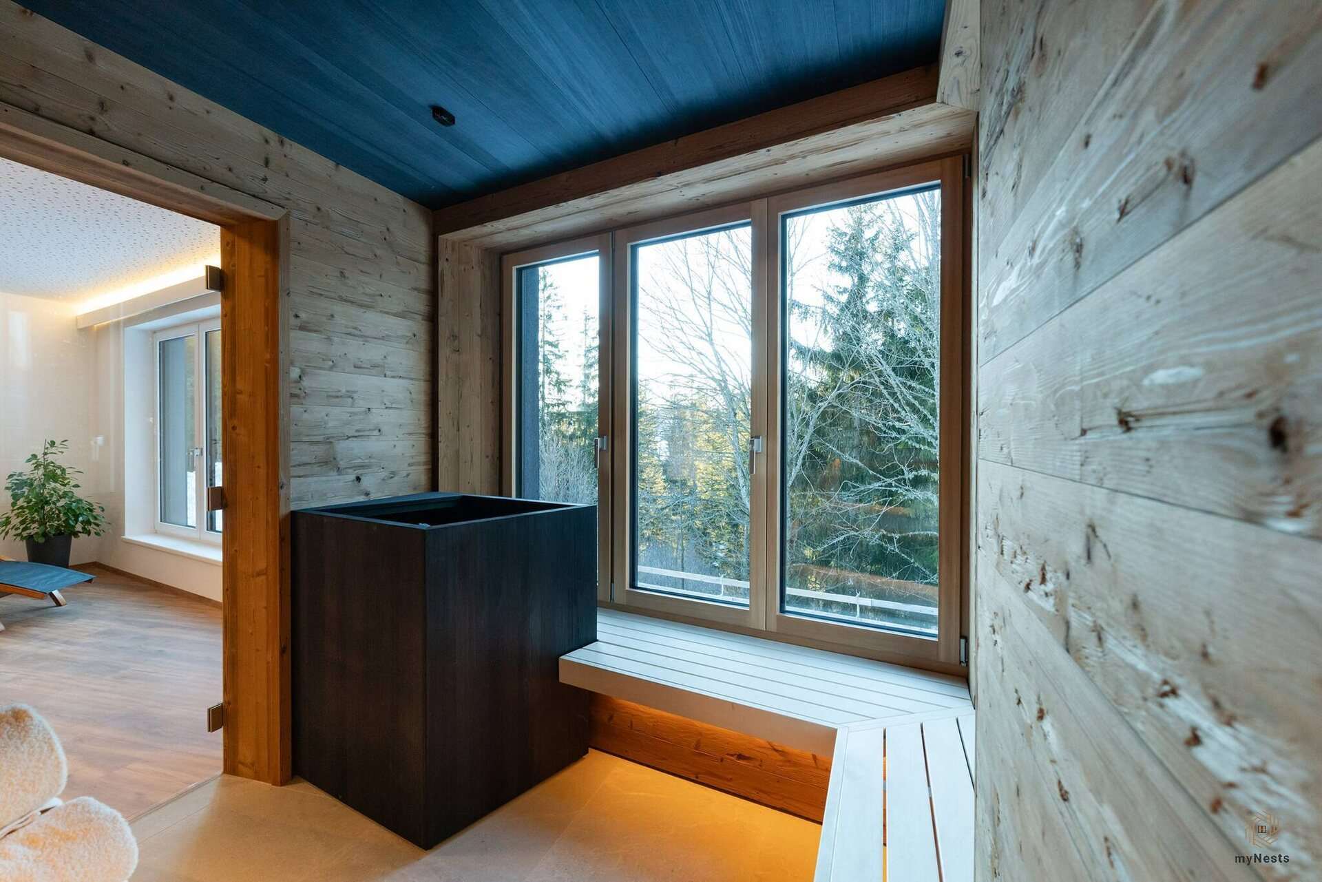 Sauna se stěnami ze starého dřeva, saunovými kamny, lavicí a velkým oknem s výhledem do lesa